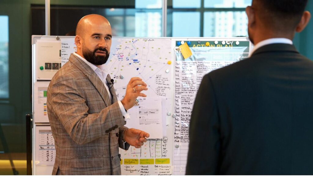 Rohollah Rohparwar erläutert vor einer Dubai-Marktkarte und einem Action-Plan-Board die Investmentstrategie im Gespräch mit einem Investor.